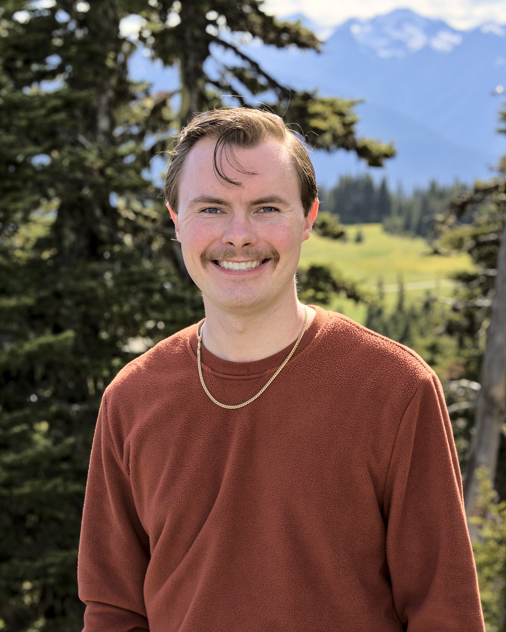 Chris Fahey smiling outdoors with evergreen trees and mountains in the background.